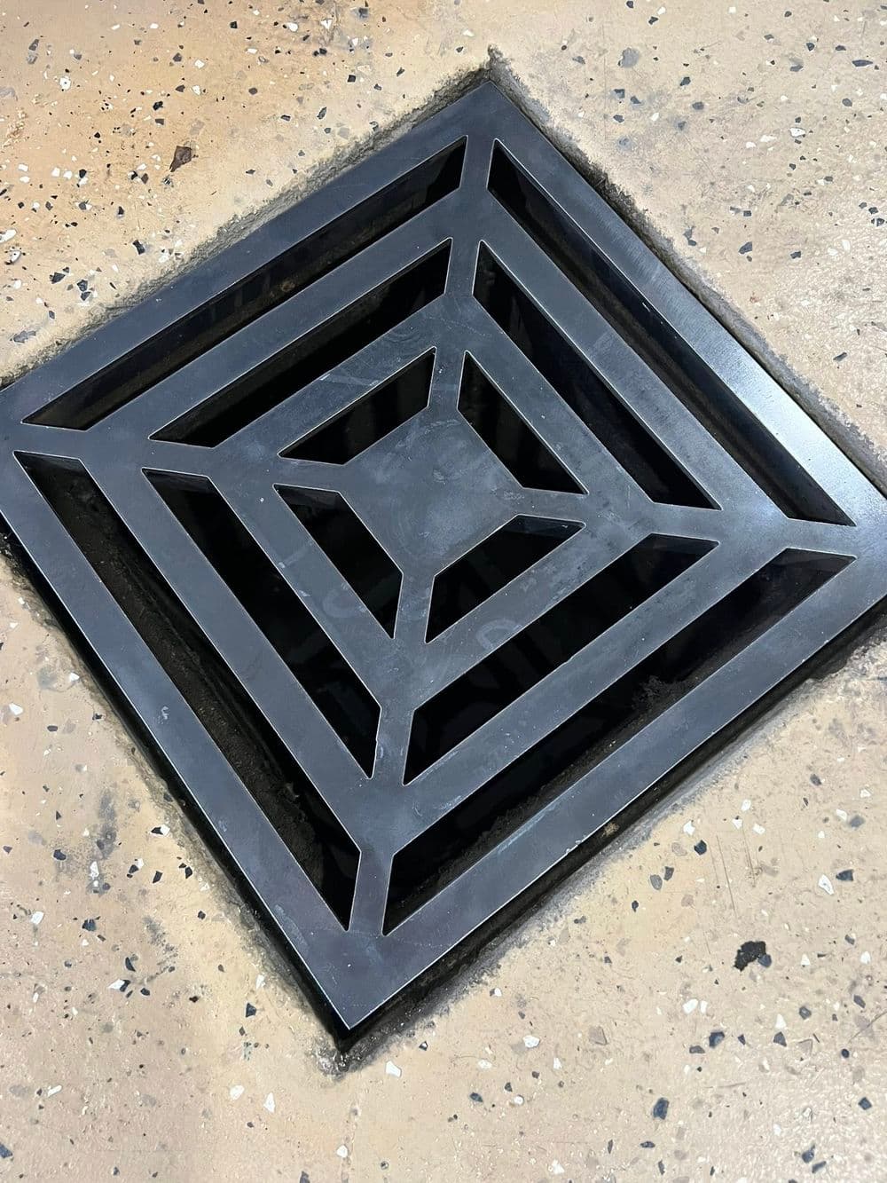 Decorative black metal drain grate with geometric pattern on a textured floor surface.