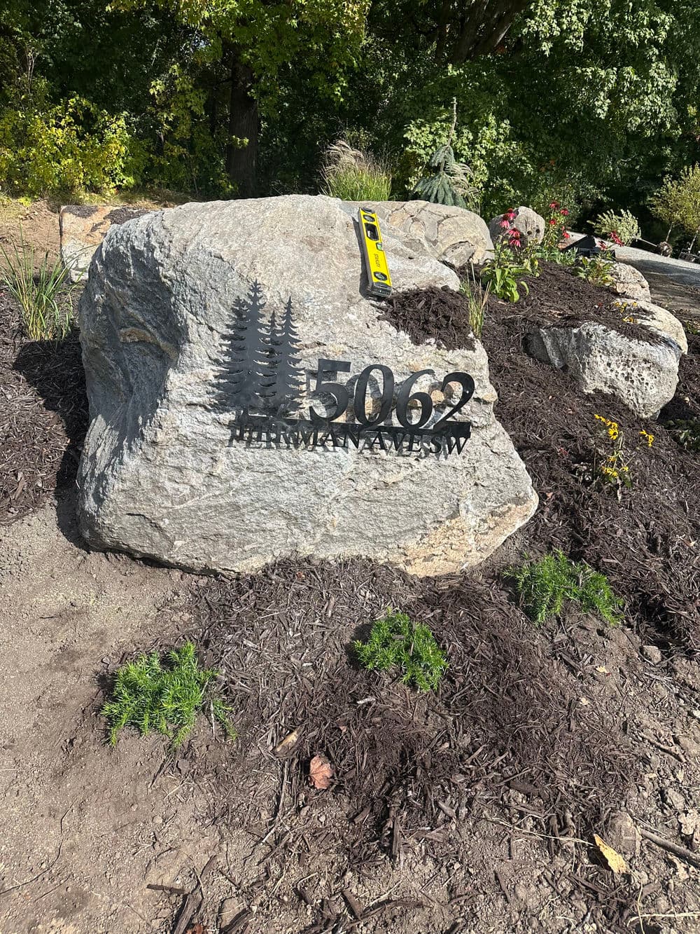 Address sign on a large rock featuring trees, located in a landscaped garden setting.